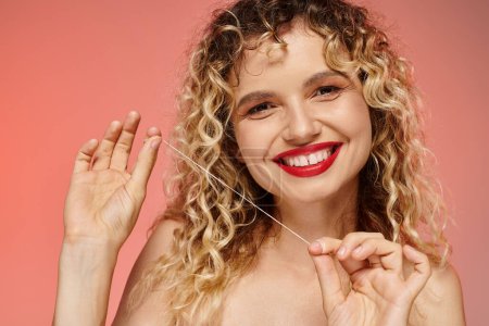 retrato de mujer feliz con cabello ondulado y labios rojos limpiando dientes con hilo dental en rosa