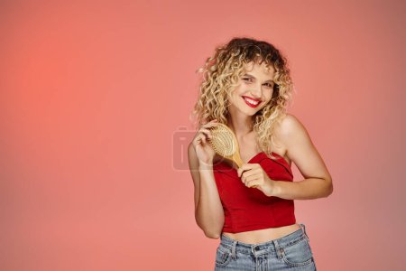 Foto de Mujer encantadora y rizada en la parte superior de moda roja posando con cepillo de pelo sobre fondo amarillo y rosa - Imagen libre de derechos