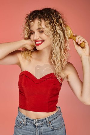 Téléchargez les photos : Femme bouclée et attrayante souriante en haut rouge brossant les cheveux ondulés sur fond de studio rose pastel - en image libre de droit