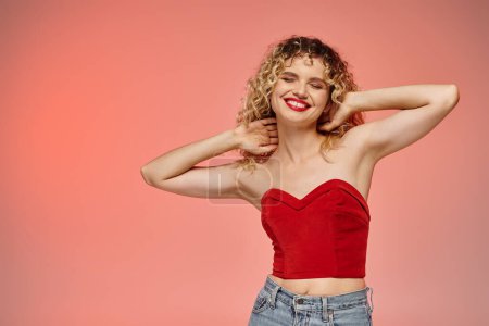 Téléchargez les photos : Femme heureuse avec des lèvres rouges et les cheveux ondulés posant en haut rouge avec les yeux fermés sur rose et jaune - en image libre de droit