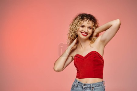 femme aux lèvres rouges et aux cheveux bouclés posant en haut rouge et souriant à la caméra sur fond pastel