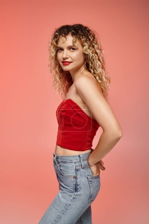 Téléchargez les photos : Femme attrayante et bouclée en haut rouge et jeans posant sur fond dégradé rose et jaune - en image libre de droit