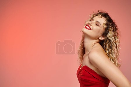 delighted woman with wavy hair posing in red top with closed eyes on pink and yellow backdrop