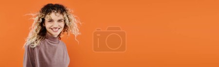 fröhliche Frau mit welligem Haar im mokafarbenen Rollkragen mit Blick in die Kamera auf orangefarbenem Hintergrund, Banner