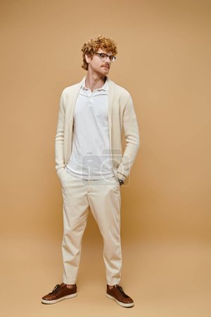 young redhead man in eyeglasses and old money style casual attire with hands in pocket on beige