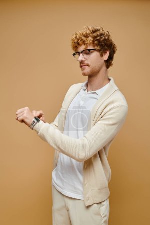 trendy redhead man in eyeglasses looking at wristwatch on beige backdrop, timeless old money fashion