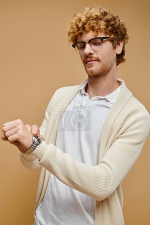 joven guapo en gafas mirando el reloj de pulsera en beige, estilo aristocrático de dinero viejo