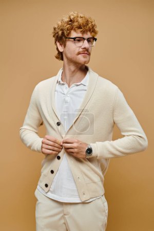 young trendy male model in eyeglasses buttoning cardigan on beige backdrop, old money fashion