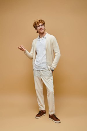 full length of elegant redhead man in eyeglasses and trendy clothes with hand in pocket on beige
