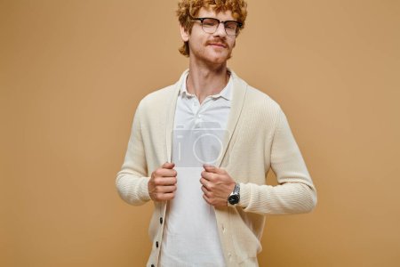 smiling redhead man in eyeglasses and old money style casual clothes looking at camera on beige