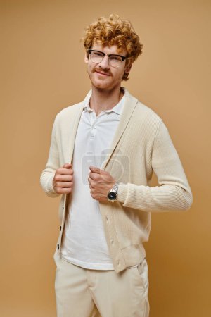 joyful redhead man in eyeglasses and old money style outfit smiling at camera on beige backdrop