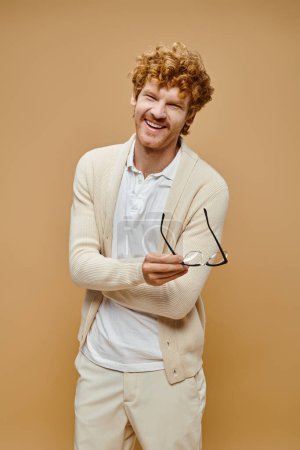 excited redhead man in light-colored clothes holding eyeglasses and laughing on beige backdrop