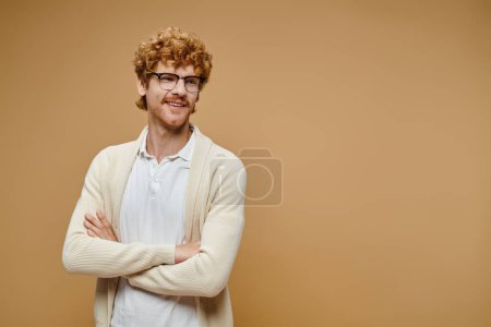 glücklicher rothaariger Mann in Brille und stylischer Kleidung posiert mit gefalteten Händen, die auf beige wegschauen