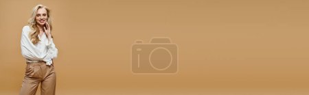 Téléchargez les photos : Femme blonde gaie en chemisier élégant et pantalon souriant à la caméra sur fond beige, bannière - en image libre de droit