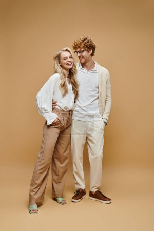 full length of cheerful couple in casual clothes smiling on beige, timeless classic fashion