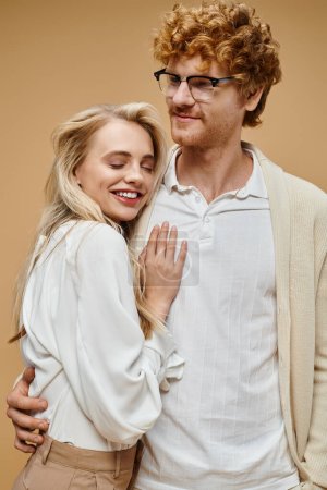 glücklicher rothaariger Mann mit Brille umarmt stylische blonde Frau lächelnd mit geschlossenen Augen auf beige
