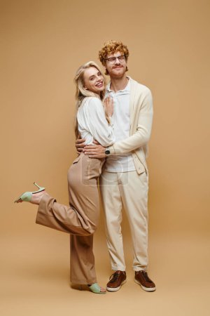 full length of carefree young models in light-colored clothes embracing on beige, classic fashion