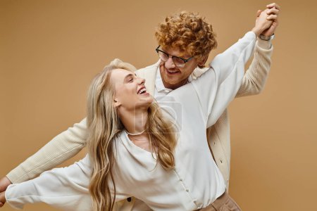 delighted fashionable couple holding hands and smiling at each other while having fun on beige