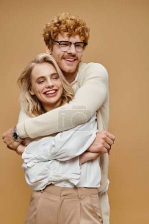 happy redhead man in eyeglasses embracing trendy blonde woman on beige, old money style couple