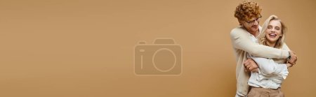 Téléchargez les photos : Homme rousse joyeux dans les lunettes embrassant femme blonde riante sur beige, bannière horizontale - en image libre de droit