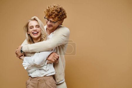 smiling redhead man in eyeglasses hugging blonde stylish woman laughing on beige, old money style