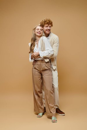 Foto de Longitud completa de alegre pareja joven en el estilo de dinero viejo atuendo casual abrazando en fondo beige - Imagen libre de derechos