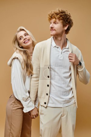 old money style, young cheerful couple in trendy outfit holding hands and looking away on beige