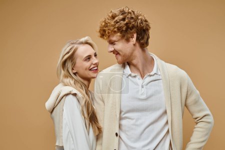 Photo for Pleased blonde woman and redhead man in old money style attire looking at each other on beige - Royalty Free Image