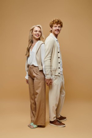full length of trendy couple in old money style clothes holding hands standing back to back on beige