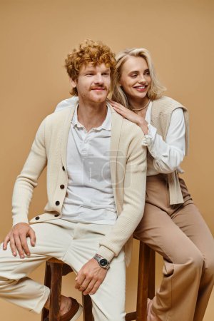 cheerful young couple in trendy clothes sitting on chairs and looking away on beige backdrop
