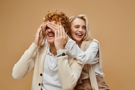 Foto de Mujer rubia juguetona cubriendo los ojos de hombre pelirrojo con estilo en color beige, moda de estilo de dinero viejo - Imagen libre de derechos