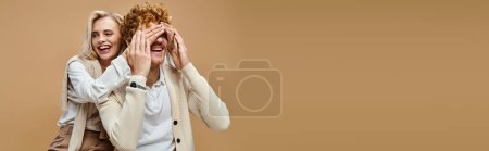 carefree blonde woman covering eyes of trendy redhead man on beige, old money style fashion, banner