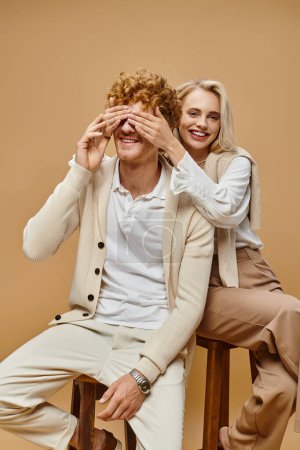 Foto de Excitada mujer rubia sentada en la silla y cubriendo los ojos del hombre pelirrojo de moda sobre fondo beige - Imagen libre de derechos