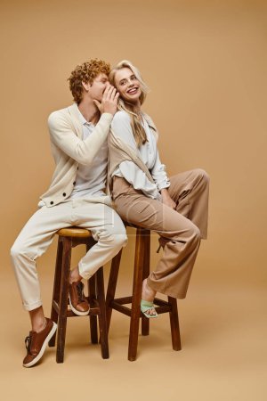 fashionable redhead man telling secret to trendy blonde woman while sitting on chairs on beige