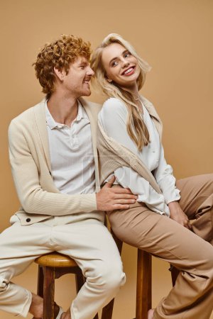 young and smiling couple in trendy casual attire sitting on chairs on grey, old money style fashion