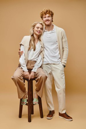 full length of redhead man embracing blonde woman sitting on chair and looking at camera on beige