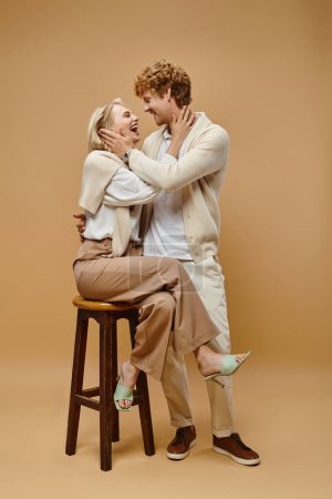 Photo for Laughing blonde woman sitting on chair and hugging young redhead man on beige, old money style - Royalty Free Image