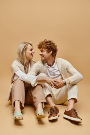 cheerful young couple in light-colored clothes sitting and smiling at each other on beige backdrop