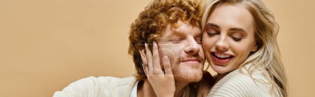 Téléchargez les photos : Femme blonde joyeux avec les yeux fermés embrassant la tête de rousse homme à la mode sur beige, bannière - en image libre de droit