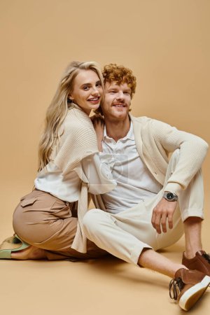 joyful and young couple in trendy old money style attire sitting on floor on beige backdrop