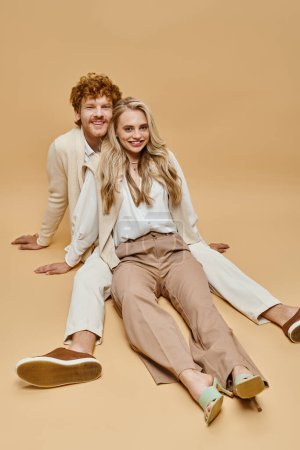 Téléchargez les photos : Couple élégant insouciant dans des vêtements de couleur claire assis et regardant la caméra sur fond beige - en image libre de droit