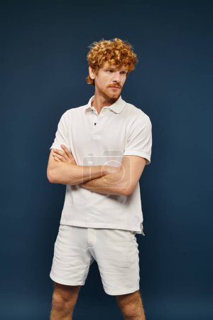 redhead young man in white polo t-shirt and shorts posing with folded arms on blue backdrop