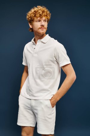 rothaariger Mann in weißem Polo-T-Shirt und kurzer Hose posiert mit den Händen in den Taschen und schaut weg auf blau