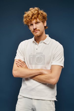 homme rousse à la mode en polo-shirt blanc regardant caméra avec bras croisés sur bleu, mode classique