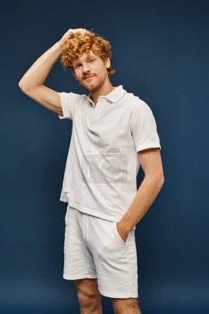 redhead man in white polo t-shirt and shorts standing with wand in pocket on blue, timeless fashion