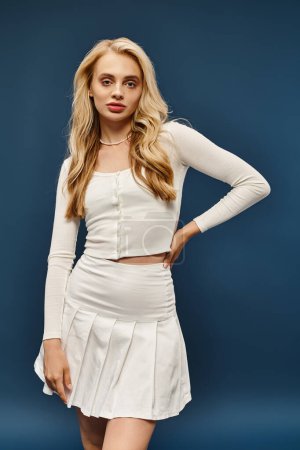 trendige blonde Frau in weißem Rock und Pullover posiert mit der Hand auf der Hüfte und blickt in die Kamera auf blau