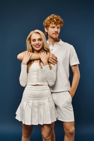 Lächelnde blonde Frau in weißer trendiger Kleidung, die in die Kamera blickt, in der Nähe eines rothaarigen Mannes, der sie auf blau umarmt