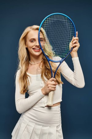 excited blonde sportive woman in white stylish attire with tennis racquet looking at camera on blue
