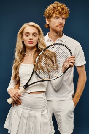 stylische blonde Frau mit Tennisschläger blickt in die Kamera in der Nähe des rothaarigen Mannes in weißen Kleidern auf blauem Grund