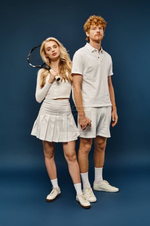 full length of blonde woman with tennis racquet near trendy redhead man in white clothes on blue